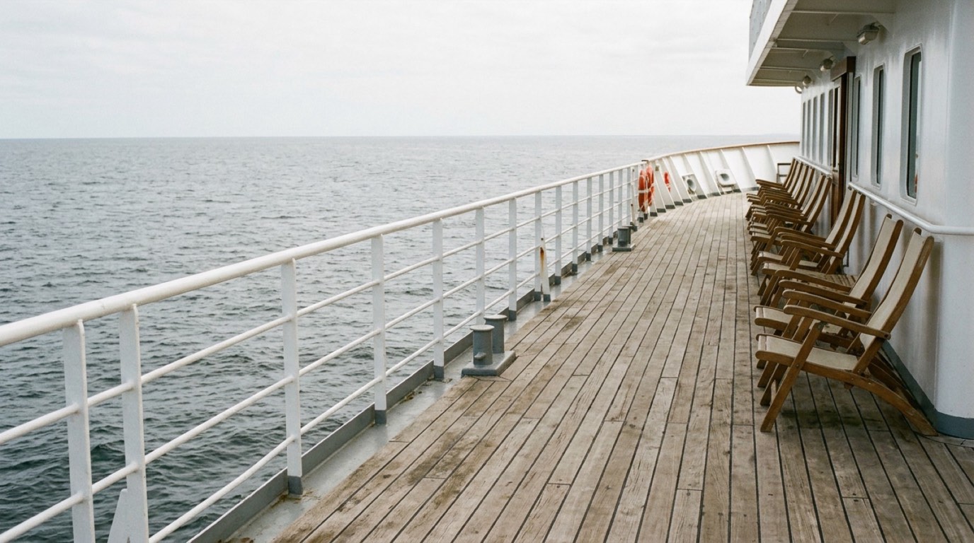 The empty upper deck of a small cruise ship at sea, with railings, teak decking and open water to the horizon under a soft overcast sky