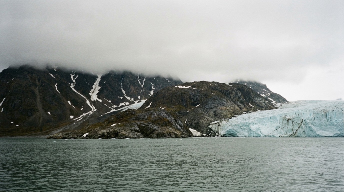 A cold northern coastline seen from the water: dark rock, streaks of snow and a pale glacier front meeting gray sea under low cloud